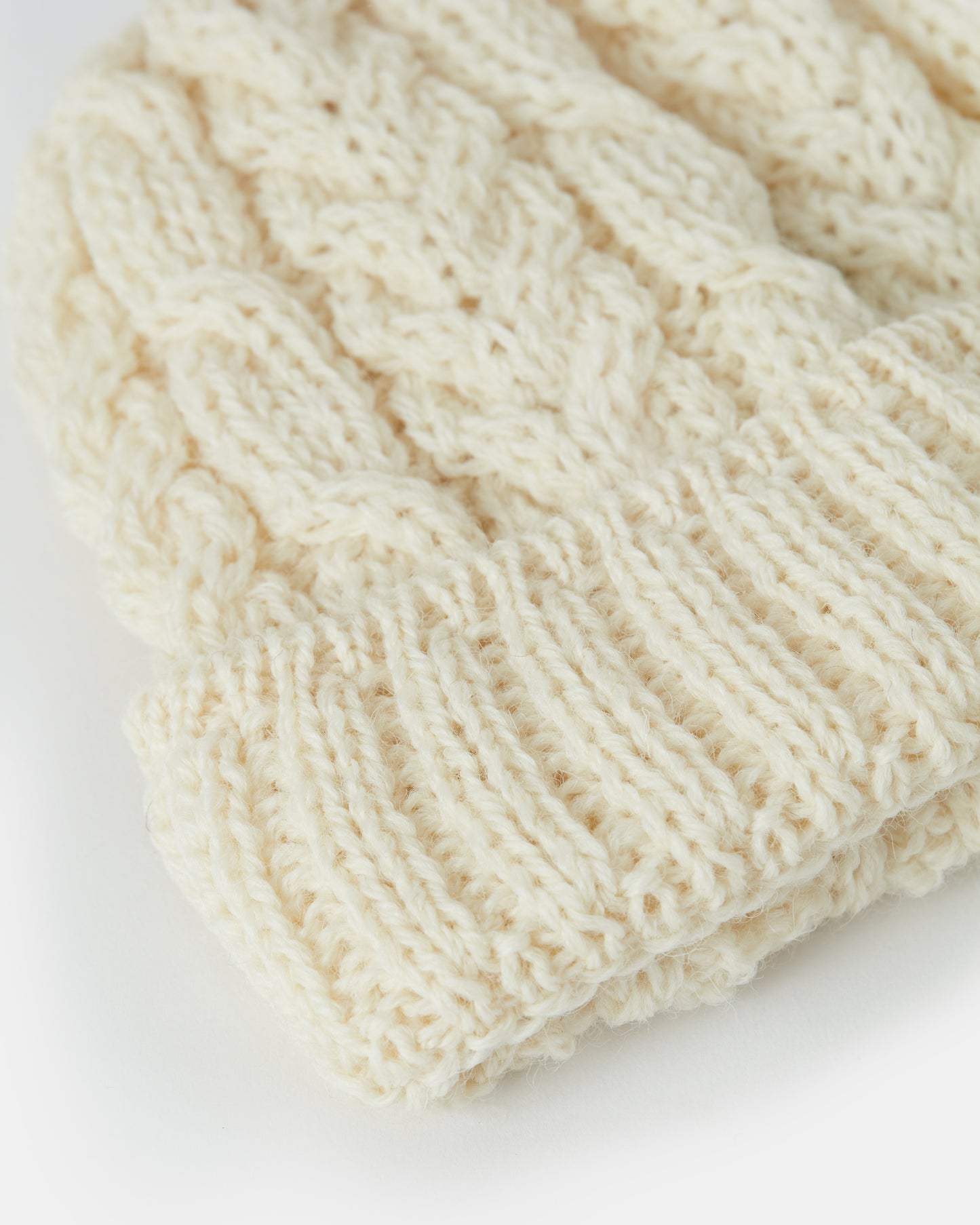 A close-up view of a cream-colored, knitted beanie hat. The beanie features a detailed cable knit pattern with a folded ribbed brim, showcasing the textured stitches of the soft fabric. This Aran Irish Knit Cable Hat - B387 by Mjölkshop.se beautifully captures the essence of traditional craftsmanship against a plain white background.