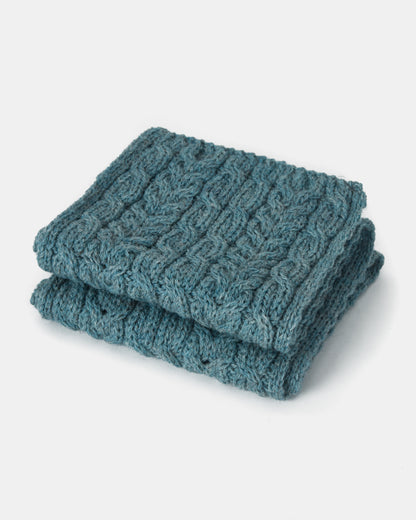 A folded, teal-colored, cable-knit Aran Irish Knit Cabel Scarf - B386 from Mjölkshop.se on a white background. Crafted from Heritage Worsted Wool, the scarf features a detailed pattern with intertwining cables, giving it a textured and cozy appearance.